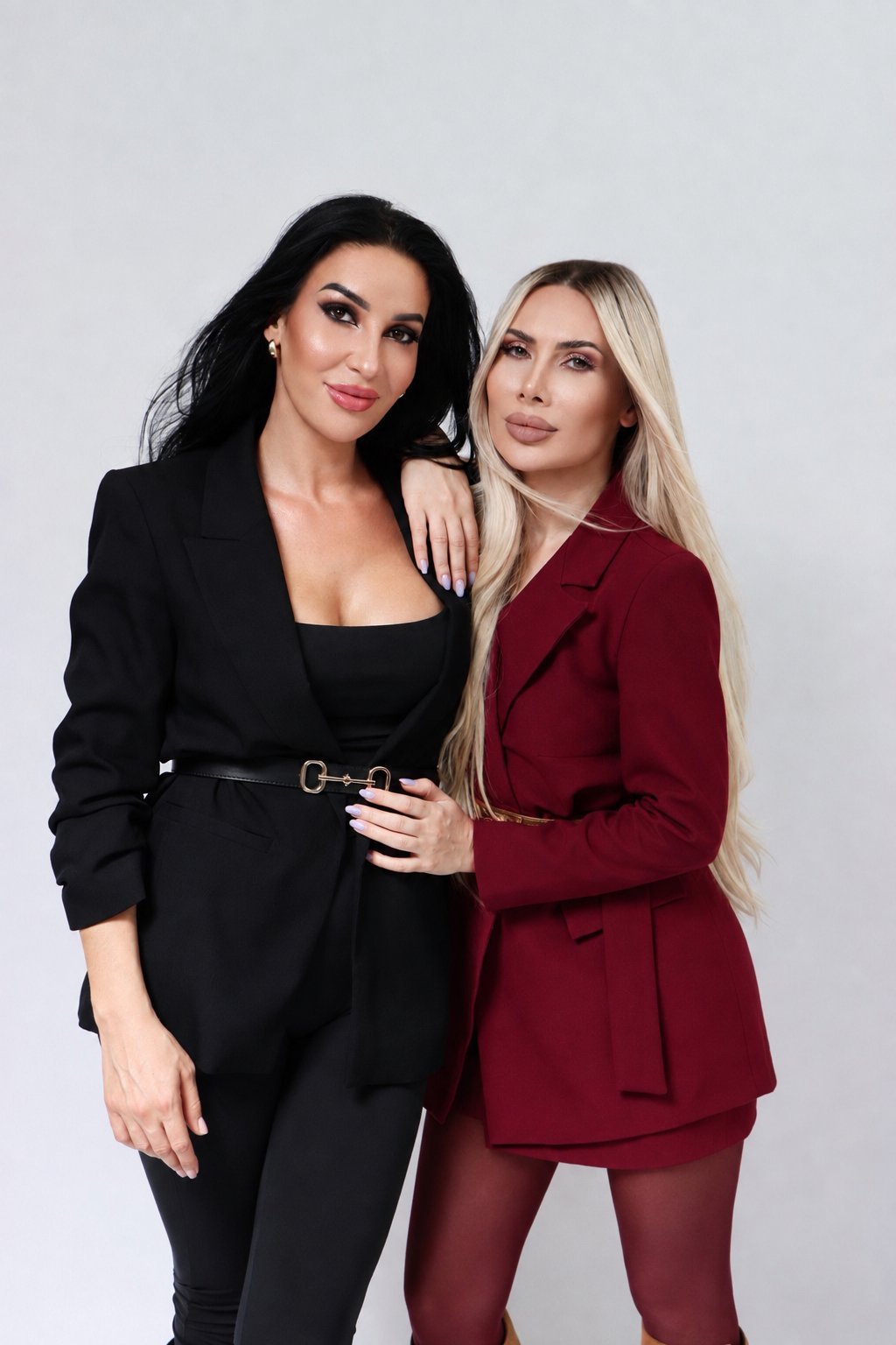 Two women posing together against a white background. Woman on left wears black blazer, woman on right wears burgundy blazer and blonde hair.